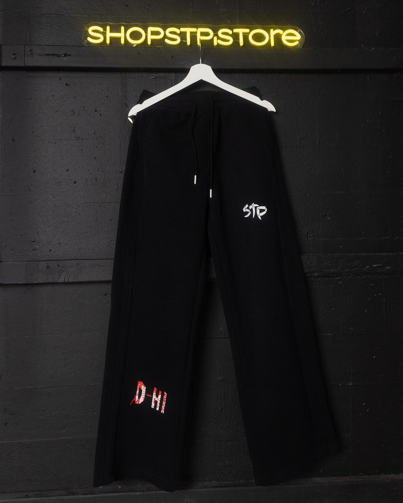 Black coat with embroidered text on a hanger against a dark wall with 'SHOPSTPSTORE' neon sign.