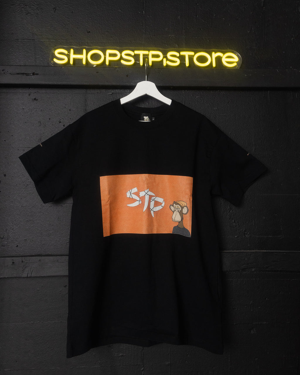 Black t-shirt with orange logo on a hanger against a dark wall with 'SHOPSTP STORE' neon sign.