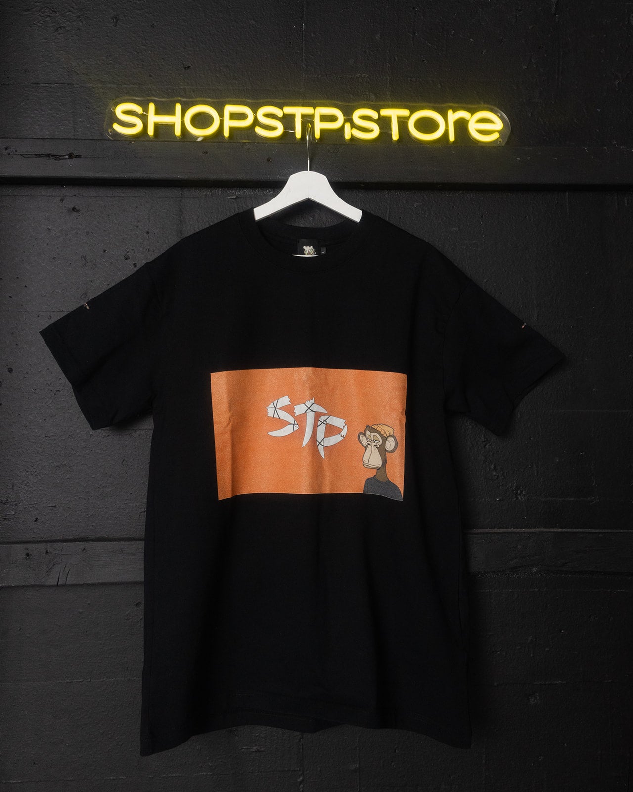 Black t-shirt with orange logo on a hanger against a dark wall with 'SHOPSTP STORE' neon sign.
