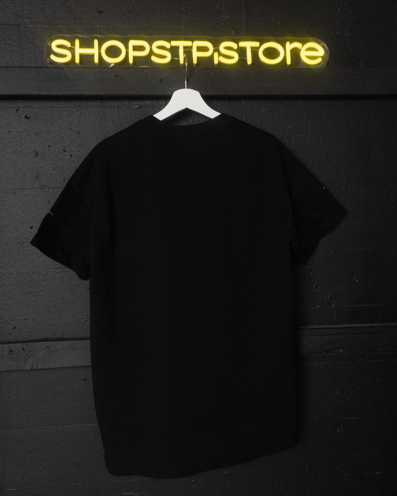Black t-shirt on a hanger with 'SHOPSTP.STORE' neon sign in the background