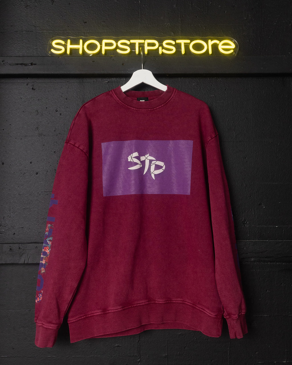 Maroon sweatshirt with a logo on a hanger against a dark wall with 'SHOPSTP.STORE' neon sign.