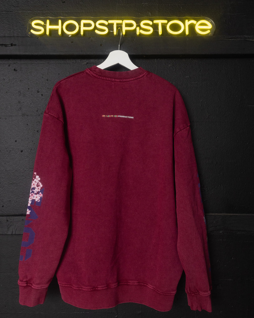 Red sweatshirt with text and graphics on a hanger against a dark background with 'SHOPSTP.STORE' neon sign.