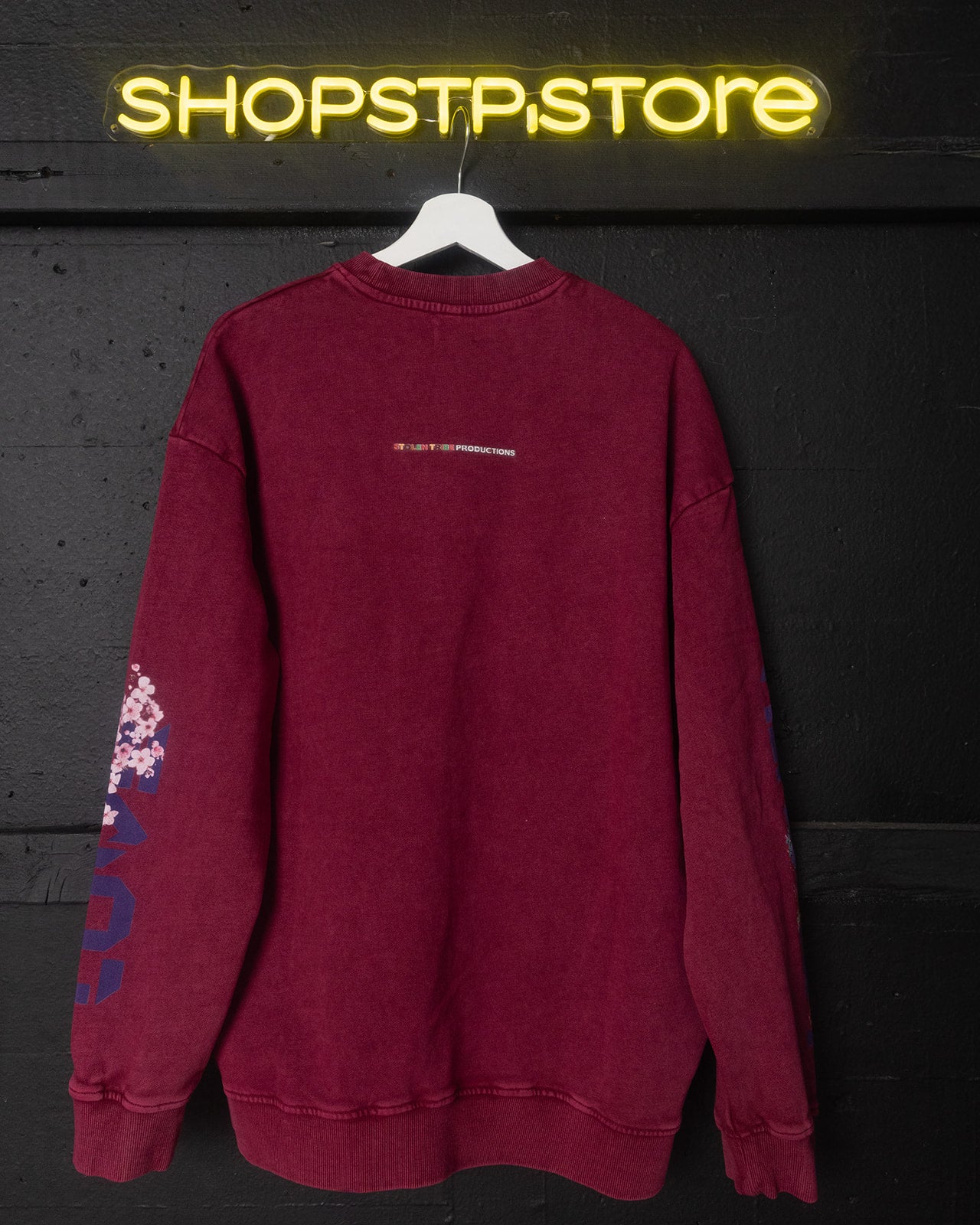 Red sweatshirt with text and graphics on a hanger against a dark background with 'SHOPSTP.STORE' neon sign.