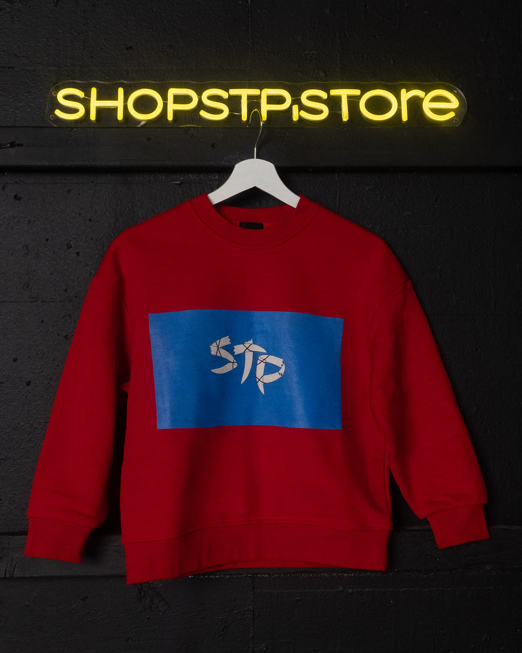 Red sweatshirt with a blue logo featuring 'STP' on a dark background with 'SHOPSTPSTORE' neon sign.