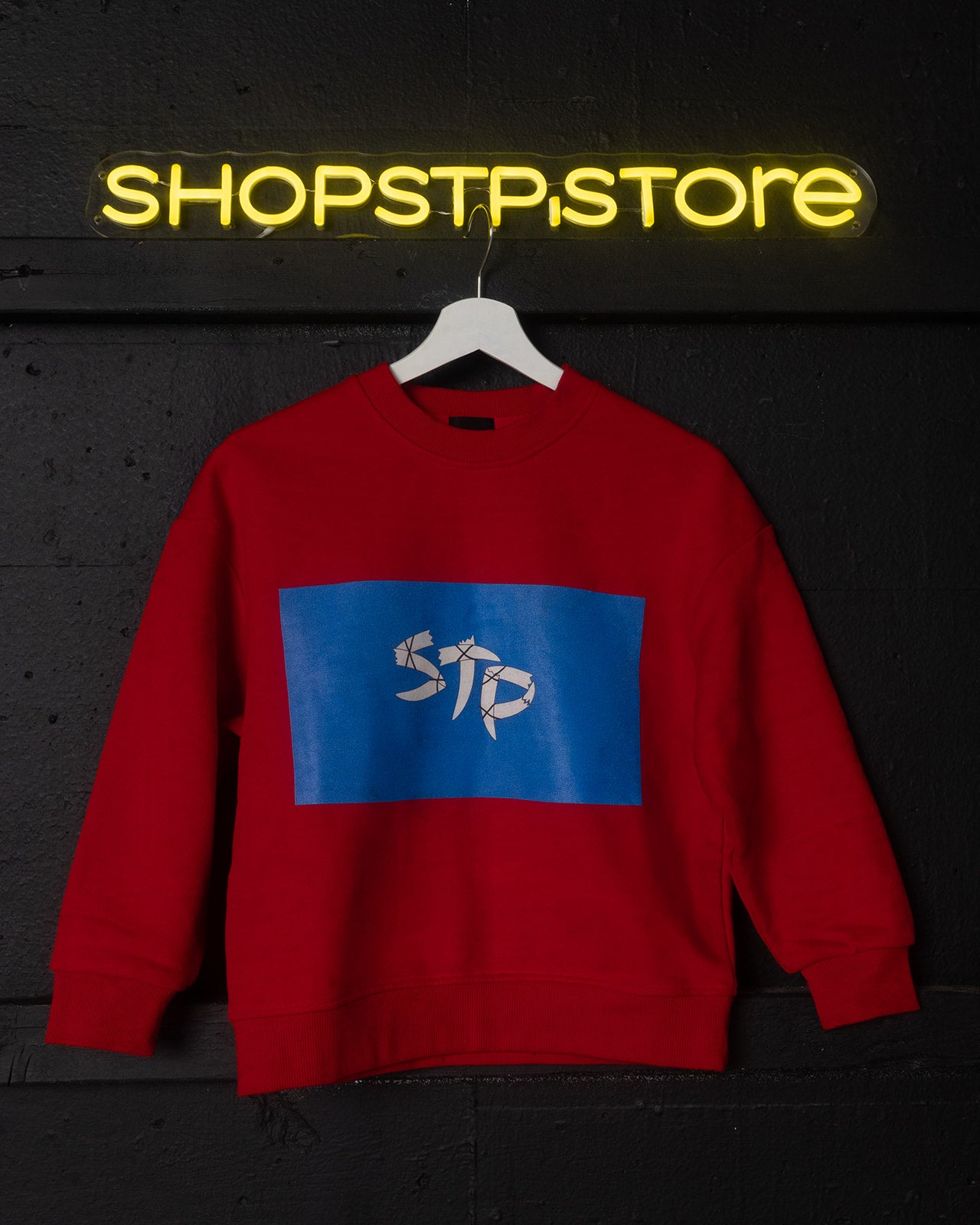 Red sweatshirt with a blue logo featuring 'STP' on a dark background with 'SHOPSTPSTORE' neon sign.