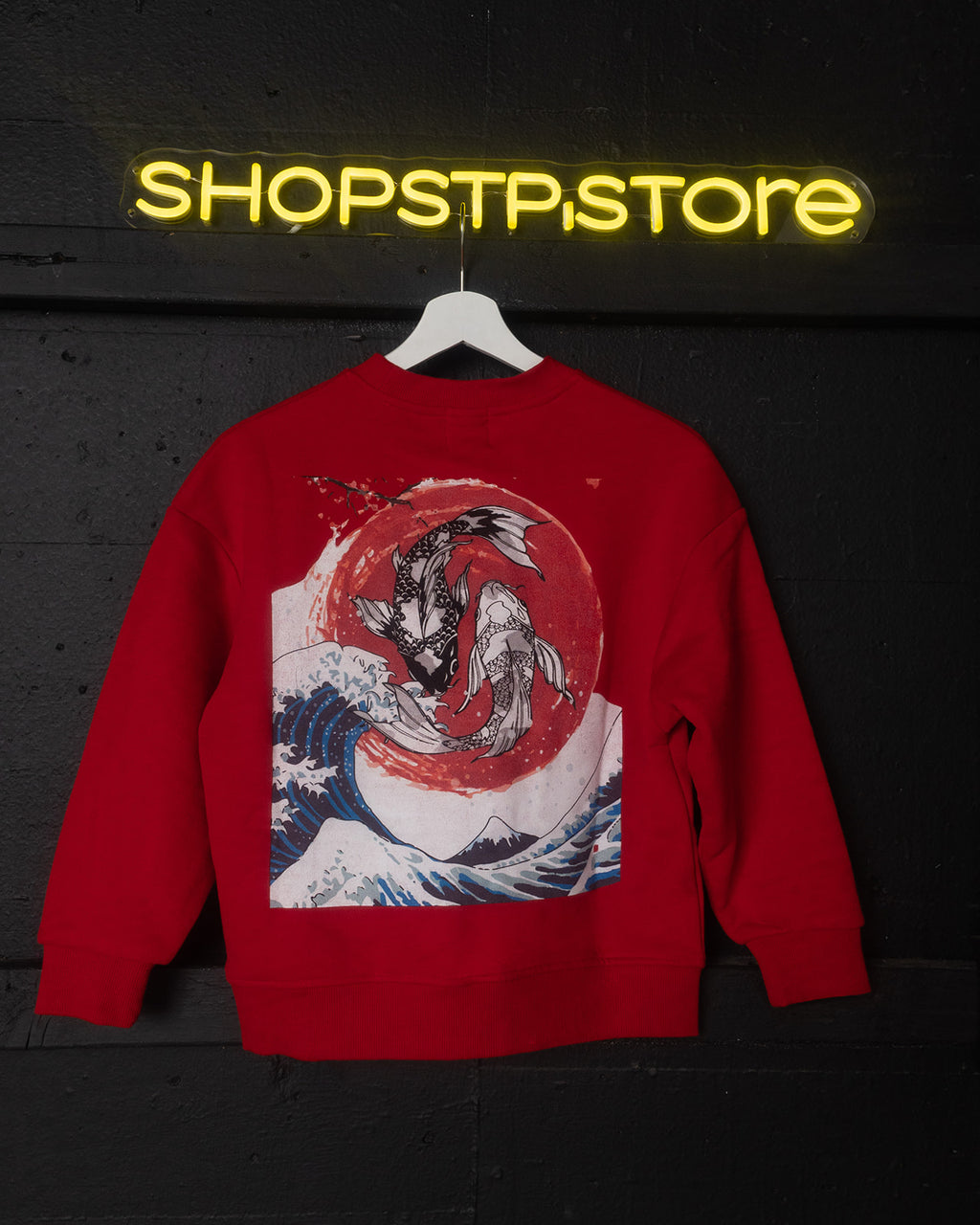 Red sweatshirt with a graphic design on a hanger against a dark background with 'Shopstpstore' neon sign.